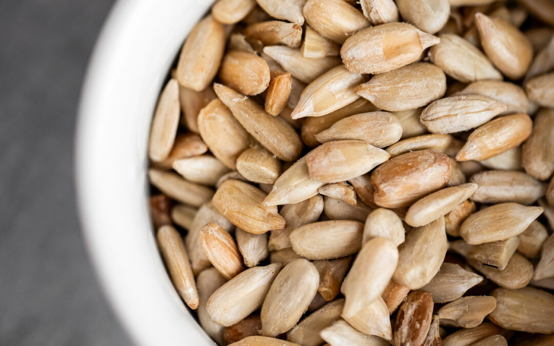 A macro shot of a white bowl filled with a pile of shelled sunflower seeds. The seeds are golden-brown and textured, captured with a shallow depth of field against a dark background.